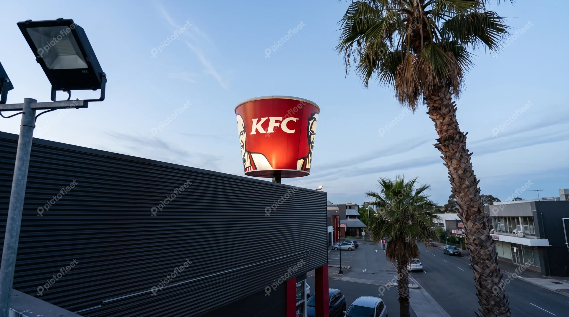 Illuminated KFC bucket sign at dusk with palm trees